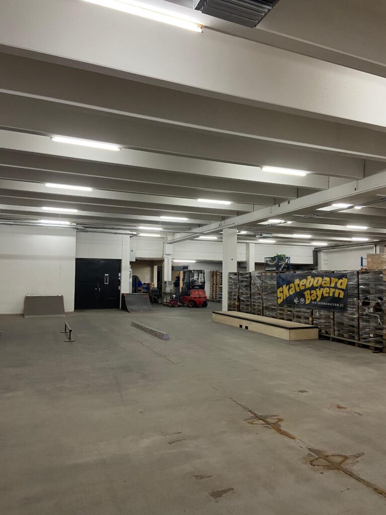 Das Bild zeigt einen weiteren Teil der Halle. Im Hintergrund stehen eine Mini-Ramp und ein Kicker an einer Wand. In der Mitte des Raumes befinden sich ein Rail, eine Curb und ein Manual Pad.