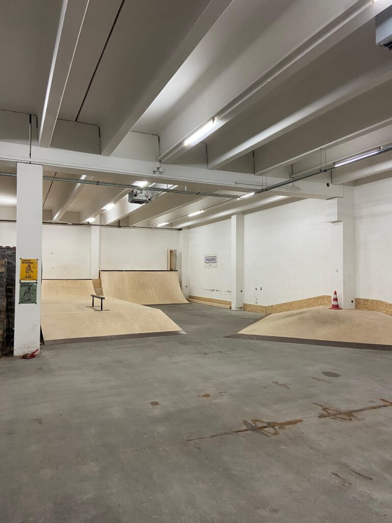 Das Bild zeigt einen Teil der Skatehalle, um die Obstacles zu präsentieren. Auf dem Bild ist eine hohe Quarterpipe in der hinteren Ecke zu sehen. An der Wand davor befindet sich eine Hip. Neben der großen Quarterpipe befindet sich eine kleinere Quarterpipe erhöht auf einem Podest, welches in einer Ramp endet und in dessen Mitte in Rail steht.