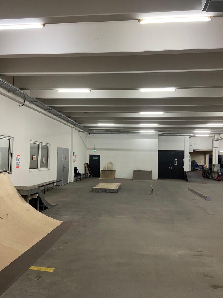 Das Bild zeigt einen leicht anderen Winkel der Halle. Das Foto ist von der mittelhohen Quarterpipe aus aufgenommen und zeigt wieder die Mini-Ramp und den Kicker an der Wand sowie Rail und Curb in der Mitte des Raumes. Auf der linken Seite ist außerdem eine kleine steile Rampe an der Wand zu sehen, die für Wall-Rides gedacht ist. Davor findet sich in der Mitte des Raums ein weiteres Manual-Pad.